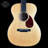 A close-up studio shot of an acoustic guitar body against a solid black background, featuring a natural wood finish, a dark reddish-brown pickguard, and a Tone Shop watermark in the upper left corner.