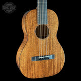 A close-up studio shot of an acoustic guitar body with a warm honey-toned wood grain finish, black neck, and silver bridge, set against a solid black background. A Tone Shop watermark is visible in the top-left corner.