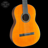 A classical acoustic guitar with a natural orange-yellow spruce top and dark brown sides, shown against a solid black background. A circular 'TONE SHOP' watermark is visible in the top-left corner.