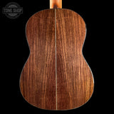 Close-up of the back of an acoustic guitar showing a rich, dark brown wood grain finish with a single central seam, set against a solid black background with a Tone Shop watermark in the top left.