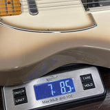 Close-up of a cream-colored electric guitar body resting on a digital scale displaying 7 lbs 0.85 oz, with the scale showing brand markings and buttons.