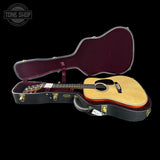 An acoustic guitar with a natural wood top and dark neck is displayed inside an open black hard case with a burgundy velvet interior, set against a solid black background. A Tone Shop watermark is visible in the top-left corner.