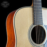 Close-up of an acoustic guitar body showing the natural wood grain top, sound hole rosette, side edge with reddish-brown varnish, and part of the dark fretboard against a black background. A Tone Shop watermark is visible in the upper left.