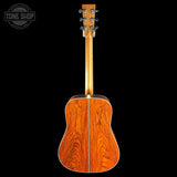 The back of an acoustic guitar with a rich, warm reddish-brown wood finish and a central binding, shown against a solid black background. A Tone Shop watermark is visible in the top-left corner.