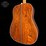 Close-up of the back of an acoustic guitar showing a rich, warm brown wood grain finish with a white binding edge, set against a solid black background. A Tone Shop watermark is visible in the top-left corner.