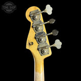 Close-up of the headstock of a Fender bass guitar with a yellow-orange sunburst finish, vintage-style metal tuners, and a Tone Shop watermark in the top left corner, set against a solid black background.
