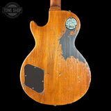The back of a vintage Gibson electric guitar with a heavily distressed orange-brown finish, showing the wood grain and dark patina. Features a circular Gibson logo badge on the upper back and a black pickguard.