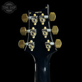 Close-up of the headstock of a black electric guitar, showing six gold tuning pegs and gold lettering that reads '10/12 STRAT ASXT' on a solid black background.