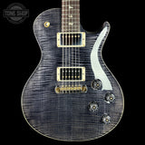 A close-up of an electric guitar body with a dark blue-gray flamed maple wood finish, gold hardware, and a white pickguard, shown against a black background with a Tone Shop watermark in the top left corner.