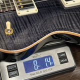 Close-up of a guitar body resting on a digital scale displaying its weight in pounds and ounces. The guitar has a dark flamed maple finish with cream-colored hardware and pickups.