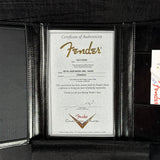 A Fender Certificate of Authenticity insert shown inside a black leather Fender instrument case. The certificate is printed on textured white paper with gold and black text and logos, including the Fender brand name and crest.