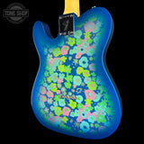 A blue electric guitar body with a vibrant, multicolored retro floral pattern in pink, green, and blue, shown against a solid black background with a Tone Shop watermark in the top left corner.