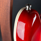 Close-up of a glossy red electric guitar body with a cream binding edge and a silver metal hardware component at the top, photographed against a dark background.