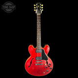 A red electric guitar with a glossy red finish, black pickguard, and gold tuning pegs, shown against a solid black background with a Tone Shop watermark in the top left corner.