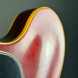 Close-up of a guitar body showing a faded red finish, a yellow binding edge, and part of a black pickguard against a dark gray background.