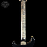 Close-up of the back of an electric guitar showing the glossy black body edge, a curved maple neck with prominent natural wood grain, and a gold-toned tuning peg, set against a solid black background.