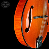 Close-up of an orange flamed maple hollow-body electric guitar body with a glittery metallic finish, featuring the f-hole, bridge, and part of the tailpiece against a black background. A Tone Shop watermark is visible in the top left corner.
