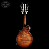 The back of a wooden mandolin with a glossy, highly figured flame maple top and six silver tuning pegs, shown against a solid black background with a Tone Shop watermark in the top left.