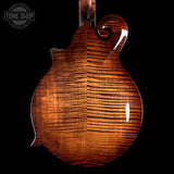 Close-up of the back of a mandolin with a glossy, highly figured brown wood finish, set against a solid black background. A Tone Shop watermark is visible in the top-left corner.