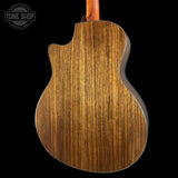 Close-up of the back of an acoustic guitar showing a rich wood grain finish, with a cutaway body shape and a dark background. A Tone Shop watermark is visible in the top left corner.