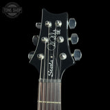 Close-up of a guitar headstock with six silver tuning pegs, white 'Steucla' and 'SE' branding, and silver strings against a solid black background.