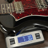 A close-up of a black electric guitar with a red glitter pickguard resting on a digital scale showing 7.30 lbs.