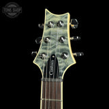 Close-up of the headstock of a custom electric guitar with a green flamed maple top, chrome tuning pegs, and a black pickguard on a black background.