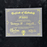 A gray Certificate of Authenticity card with gold foil lettering and border, placed on a black textured background. The card is signed by Elliot Rowe and dated 8/1/26, certifying a Rock N' Roll Rails custom instrument with model Revenge Jr.