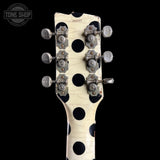 Close-up of the headstock of a vintage electric guitar with a cream-colored finish, black polka dots, and silver tuning pegs, shown against a solid black background.