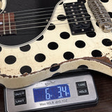 A close-up of a white electric guitar with large black polka dots and a heavily distressed, cracked vintage finish, shown resting on a digital scale displaying 6.34 oz.