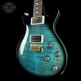 A close-up of an electric guitar body with a vibrant teal-blue flame finish and a dark wood neck, shown against a solid black background. A circular 'TONE SHOP' watermark is visible in the top-left corner.