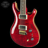 A red electric guitar with a glossy flamed maple top, gold hardware, and a black pickguard, shown against a solid black background with a Tone Shop watermark in the top left corner.