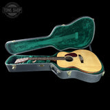 An acoustic guitar with a natural wood top and dark neck, displayed inside an open gray hard case with a green velvet interior, set against a solid black background.