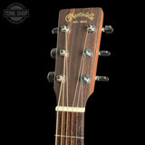 Close-up of the headstock of a Martin & Co acoustic guitar against a black background, showing the dark wood grain, gold lettering, and silver tuning pegs.