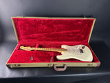 An electric guitar with a cream-colored body and maple neck, resting inside an open vintage hard case lined with red velvet.