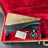 A guitar pickup kit displayed inside an open red velvet-lined case, including a gold-toned metal plate, a black leather strap, a silver metal plate, and a black circular component with a compass-like emblem.