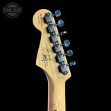 Close-up of the headstock of a Fender electric guitar in natural maple wood, featuring six silver tuning pegs and a signed autograph on the backplate, set against a solid black background.
