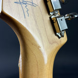 Close-up of the headstock of an electric guitar, showing a light natural wood finish with a blue signature inlaid near the top and silver tuning pegs against a dark background.