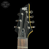 Close-up of the headstock of an SGT electric guitar against a solid black background, showing the black glossy headstock, six silver tuning pegs, strings, and a yellow edge outline.