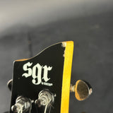 Close-up of a guitar headstock with a black face, white gothic-style 'sgr' logo, gold binding, and tuning pegs against a dark blurred background.