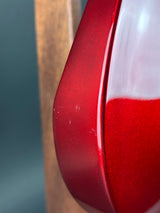 Close-up of a glossy red guitar body with a white headstock partially visible, photographed against a blurred gray background.
