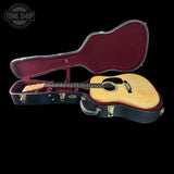 An acoustic guitar with a natural wood top and black headstock, displayed inside an open black hard case with a burgundy velvet interior, set against a solid black background.