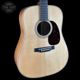 An acoustic guitar with a natural wood finish and a dark tortoiseshell pickguard, shown against a solid black background with a Tone Shop logo in the top-left corner.
