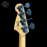 Close-up of the headstock of an electric guitar, showing a natural maple wood neck with four black tuning pegs and a black signature logo, set against a solid black background.