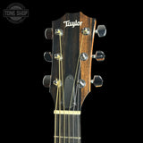 Close-up of the headstock of a Taylor acoustic guitar against a black background, showing the brand name Taylor in white script, six tuning pegs, and six strings. A Tone Shop watermark is visible in the top left corner.