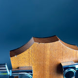 Close-up of the headstock of a wooden stringed instrument, showing the wood grain, edge profile, and part of the tuning pegs against a dark blue background.