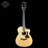 A Taylor acoustic-electric guitar with a natural wood finish and dark cutaway body, shown against a solid black background with a Tone Shop watermark in the top-left corner.