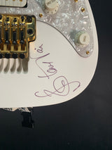 Close-up of a white electric guitar body with gold hardware, a clear pearlescent pickguard, and a purple signature on the side, photographed against a dark background.