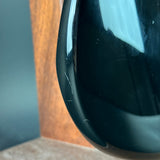 Close-up of a glossy black teardrop-shaped decorative object, likely a vase or sculpture, showing minor surface scratches and reflections, set against a blurred background with a wooden panel and gray wall.