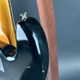Close-up detail of a glossy black electric guitar body with a gold-toned sunburst edge and part of a brown wooden stand visible against a dark gray background.